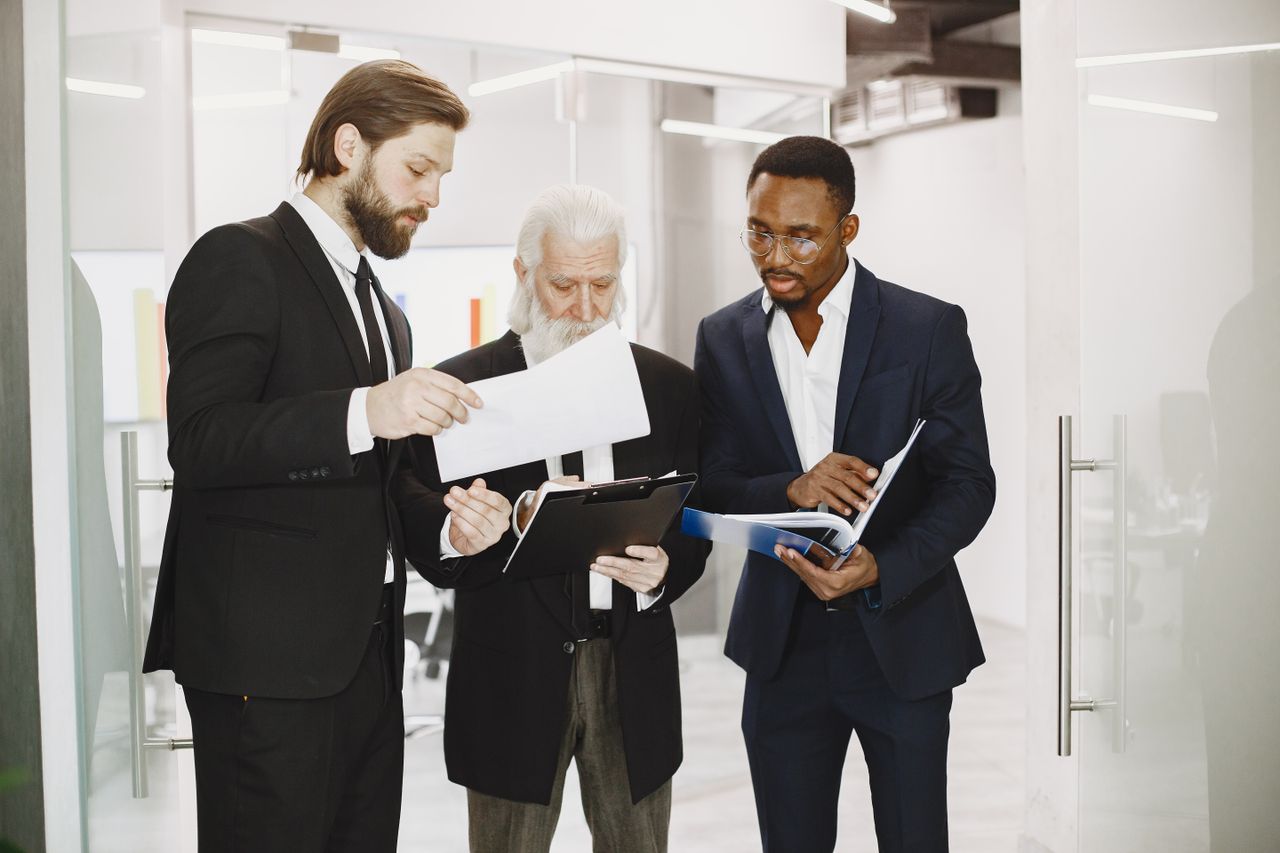 Three diverse businessmen in suits collaborate over documents in a modern office setting.